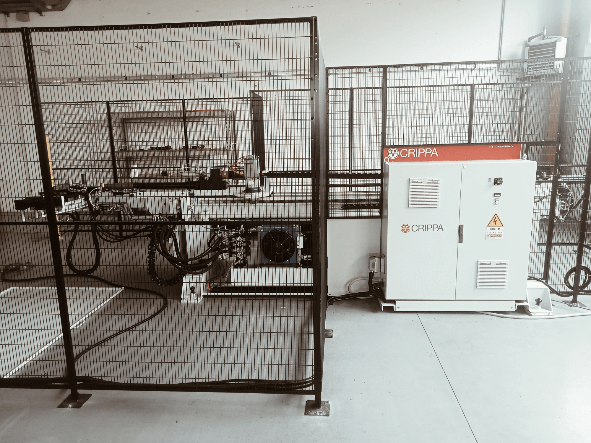 Weiße CRIPPA-Maschine hinter einem Sicherheitskäfig, mit sichtbaren Schläuchen und Komponenten, Seitenansicht, Industrieumgebung.