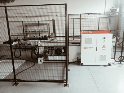 Weiße CRIPPA-Maschine hinter einem Sicherheitskäfig, mit sichtbaren Schläuchen und Komponenten, Seitenansicht, Industrieumgebung.