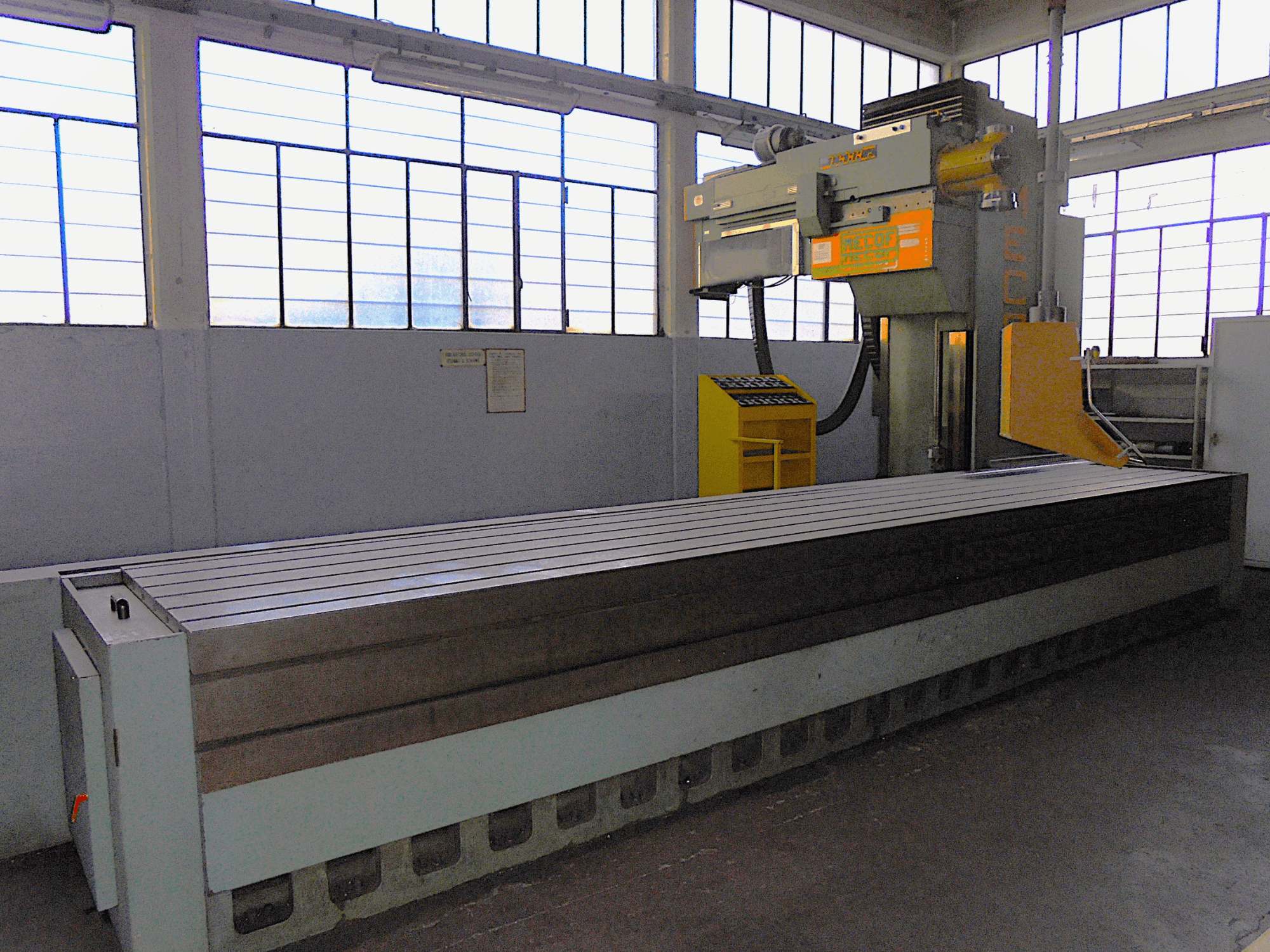 Vertikale Fräsmaschine (Marke unbekannt) in einer Fabrik, Seitenansicht, mit großem Tisch mit gerillter Oberfläche und Bedienfeld.