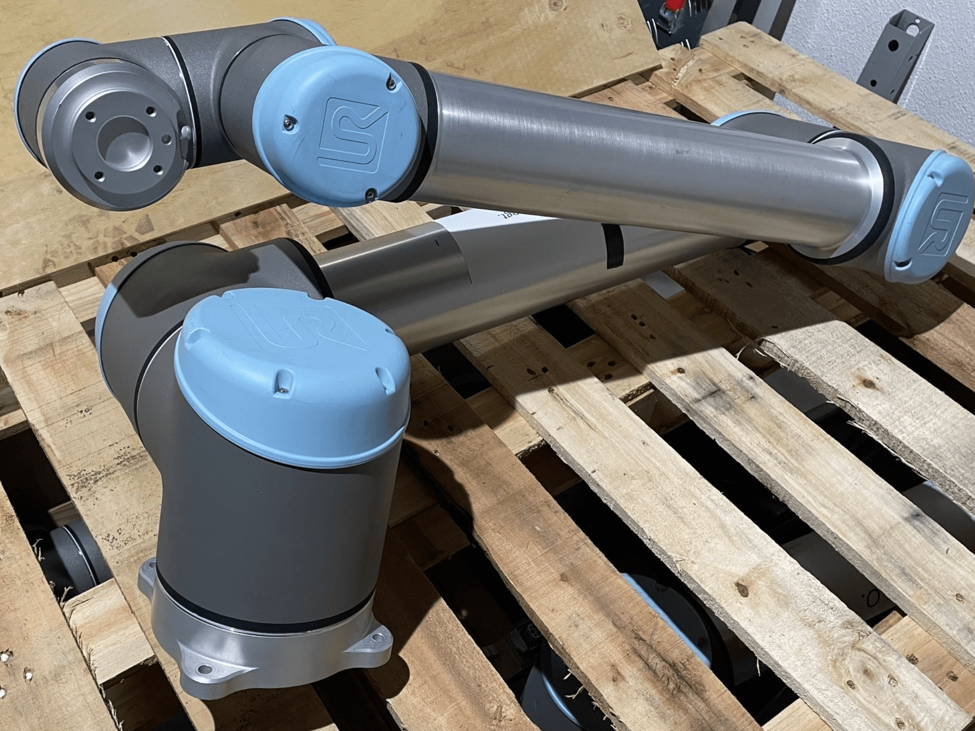 Silberner Roboterarm mit blauen Endkappen, Vorderansicht, mit Gelenken und mechanischen Details, auf einer Holzpalette ruhend.