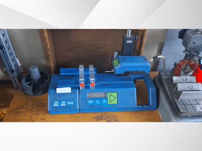 Blauer Hommel T 1000 Drucker und T 1 010 Tester mit roten Tasten, von vorne gesehen auf einer Werkbank mit Metallblöcken.