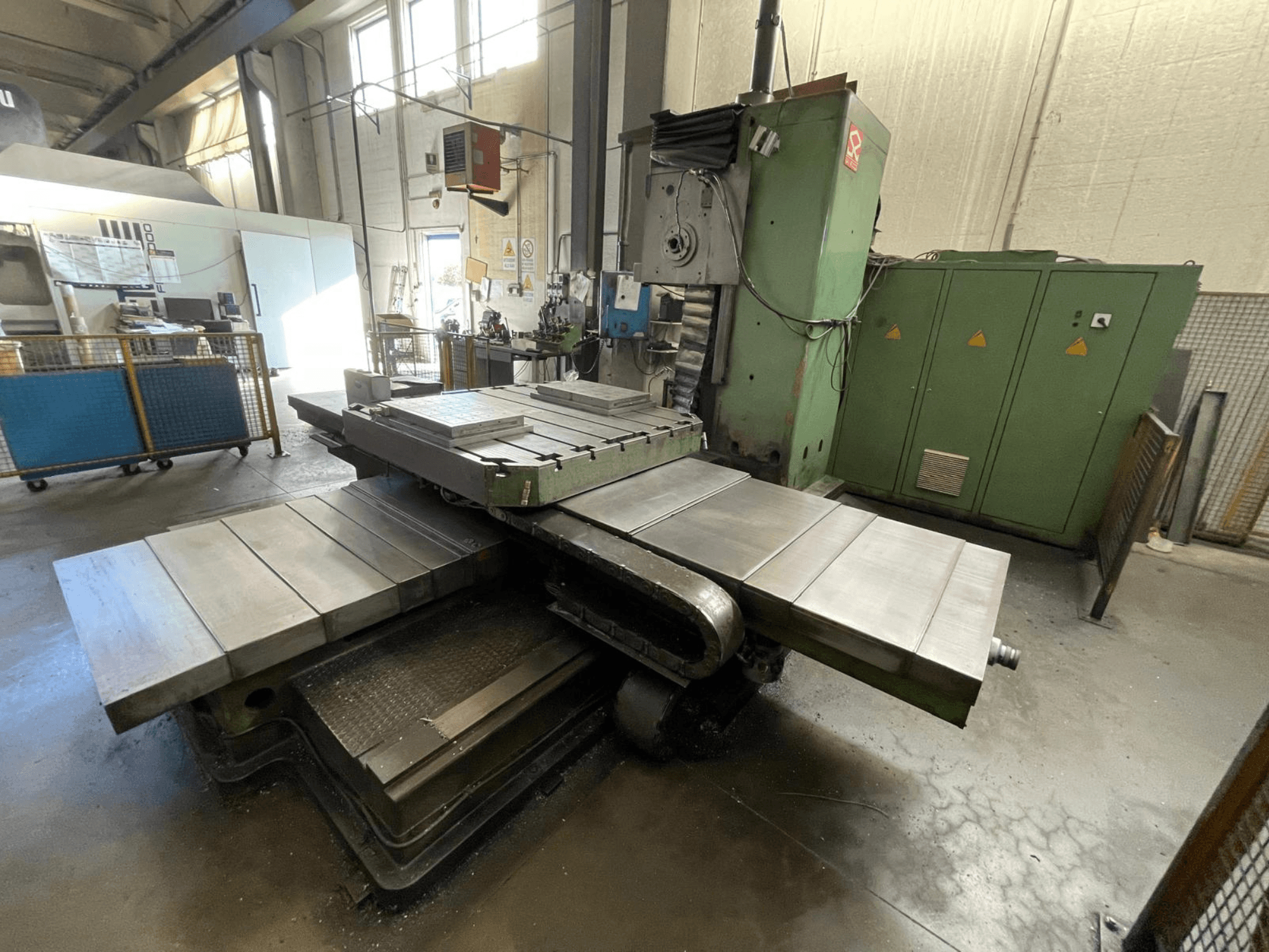 Große industrielle Fräsmaschine mit grünem Gehäuse, Seitenansicht, mit mehreren ebenen Flächen und Lagerschränken im Hintergrund.