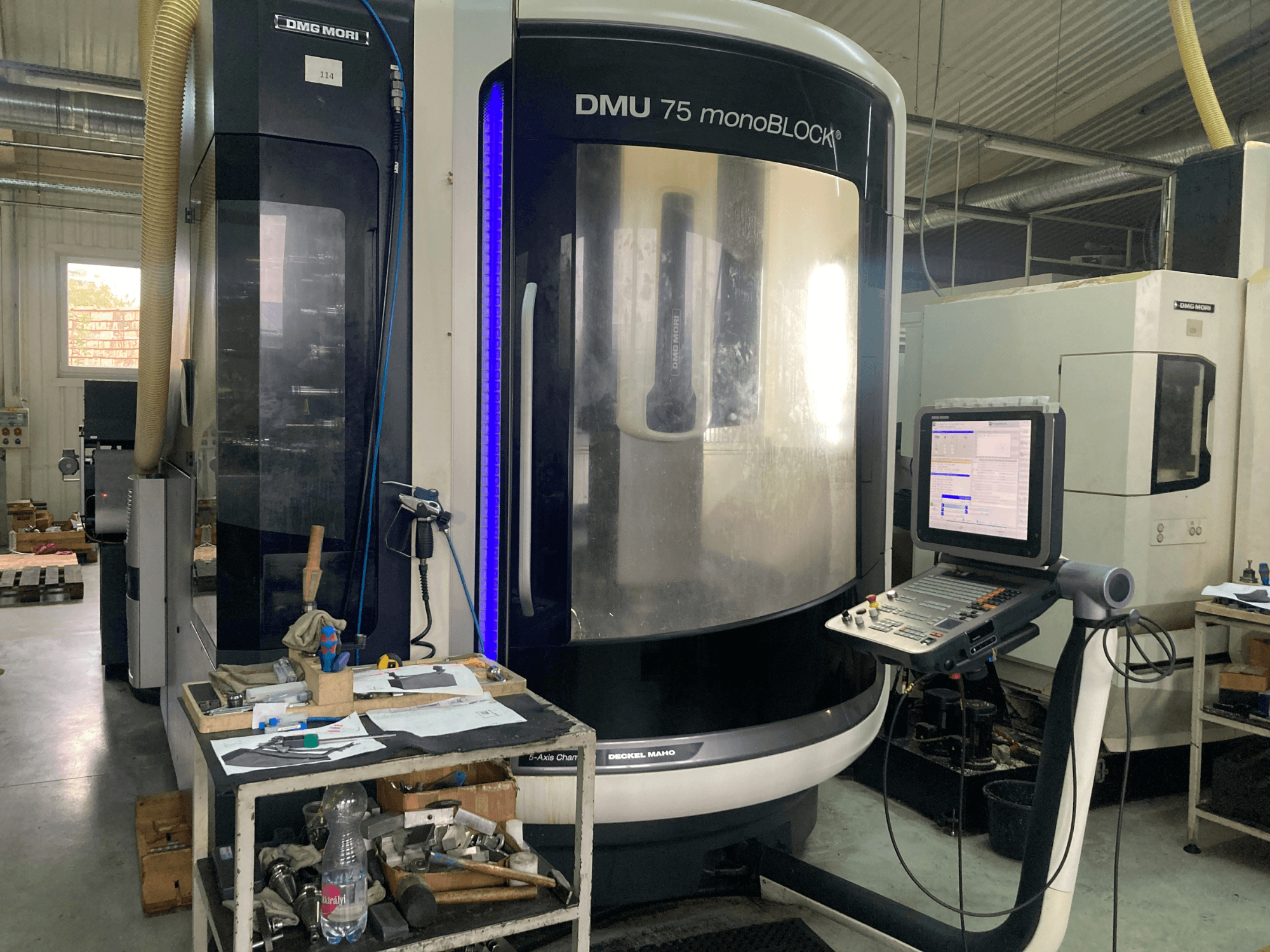 DMG MORI DMU 75 monoBLOCK® CNC-Maschine, Vorderansicht, mit Bedienfeld, Werkzeugen und einem Arbeitsbereich mit Materialien.