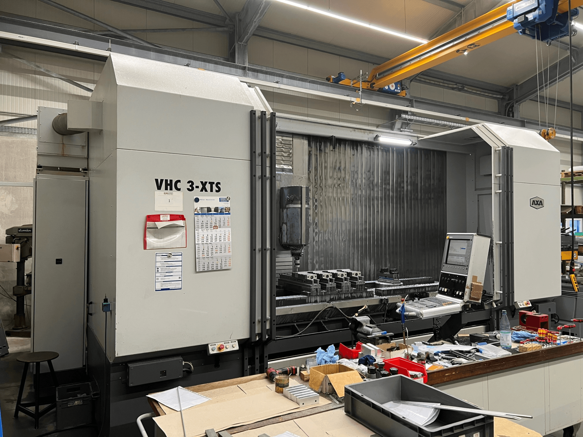 CNC-Maschine AXA VHC 3-XTS in der Seitenansicht, mit Bedienpult und einem mit Werkzeugen und Bauteilen überfüllten Arbeitsbereich.