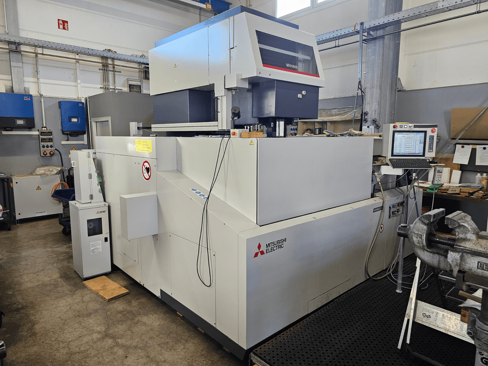 Mitsubishi MV4800 Drahterodiermaschine in einer Werkstatt, Seitenansicht mit Bedienfeld, Sicherheitsschildern und umliegenden Geräten.