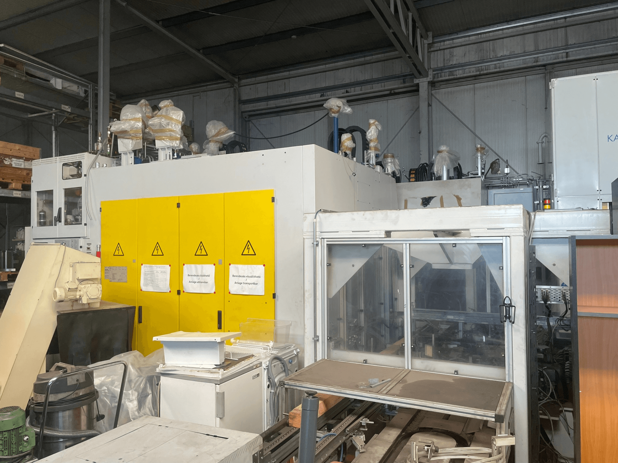 Industriemaschine mit gelbem Bedienfeld, Seitenansicht, mit Warnschildern und Kunststoffabdeckungen, in einer Lagerhalle.