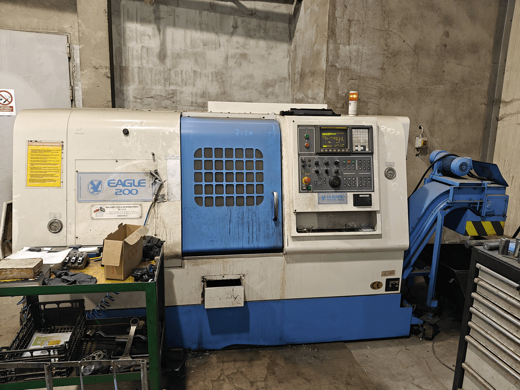 Eagle 200 CNC-Maschine, Frontansicht mit Bedienfeld, verschiedene Werkzeuge auf einem grünen Wagen. Sichtbare Abnutzung und Betriebsleuchten.