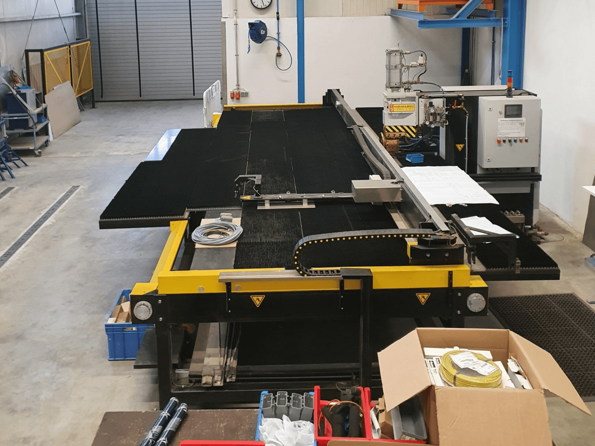 Industrielle Schneidemaschine mit gelben und schwarzen Merkmalen, Vorderansicht, mit Bedienelementen und Zubehör in einer Fabrikumgebung.