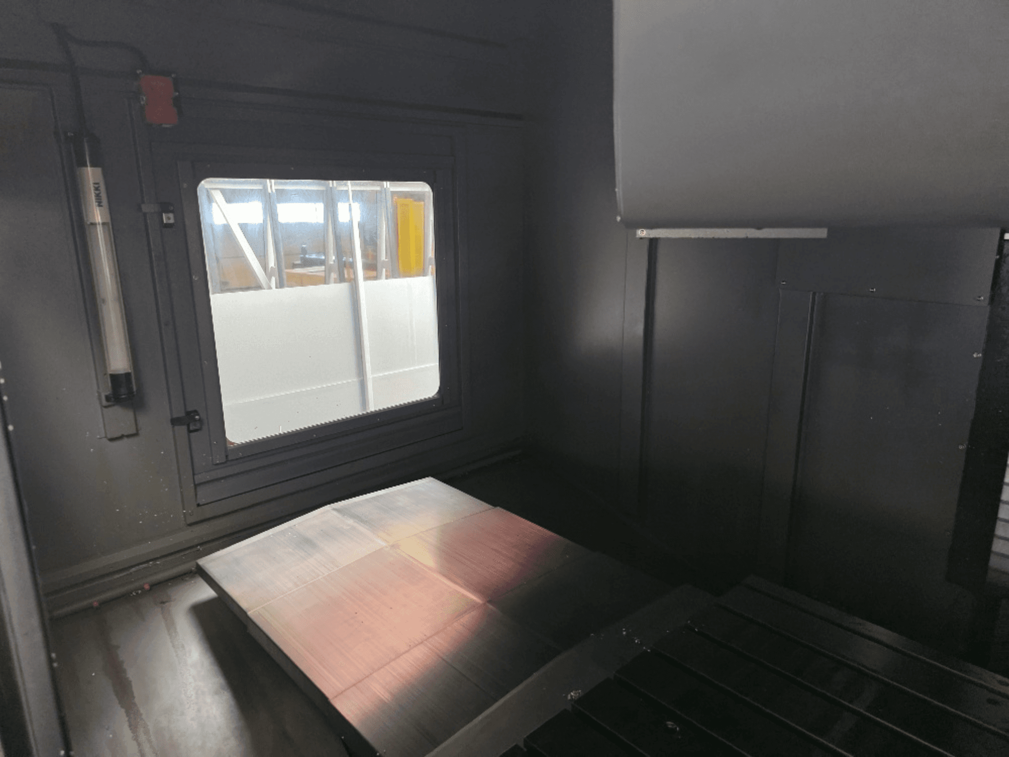 Innenansicht eines Maschinengehäuses mit einer polierten Metallplatte, einem Seitenfenster und minimaler Beleuchtung. Keine Marke sichtbar.