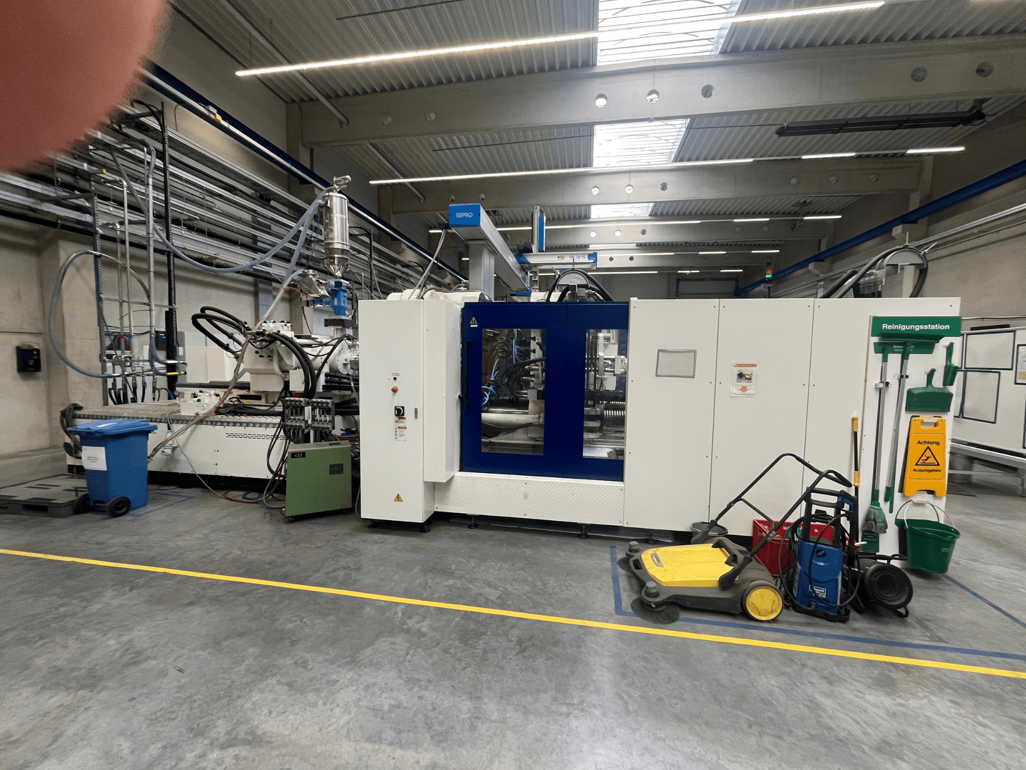 Industriemaschine in einer Fabrik, Vorderansicht, mit blauen und weißen Platten, Schläuchen und Reinigungsgeräten in der Nähe.