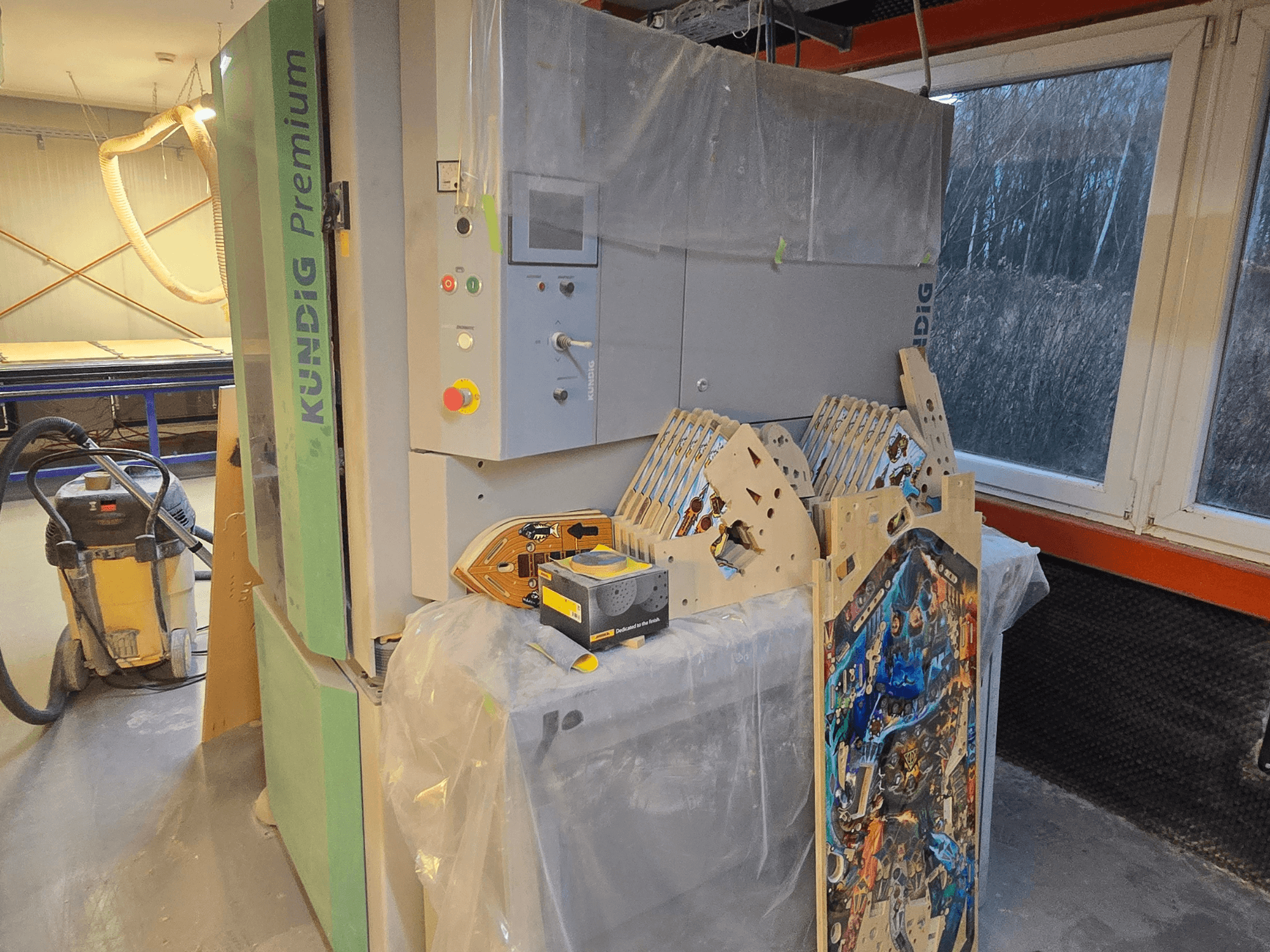 Kundig Premium-1 1100-C Bandschleifmaschine in der Werkstatt, mit Bedienfeld und Holzteilen, Seitenansicht, mit Staubabsaugung im Hintergrund.