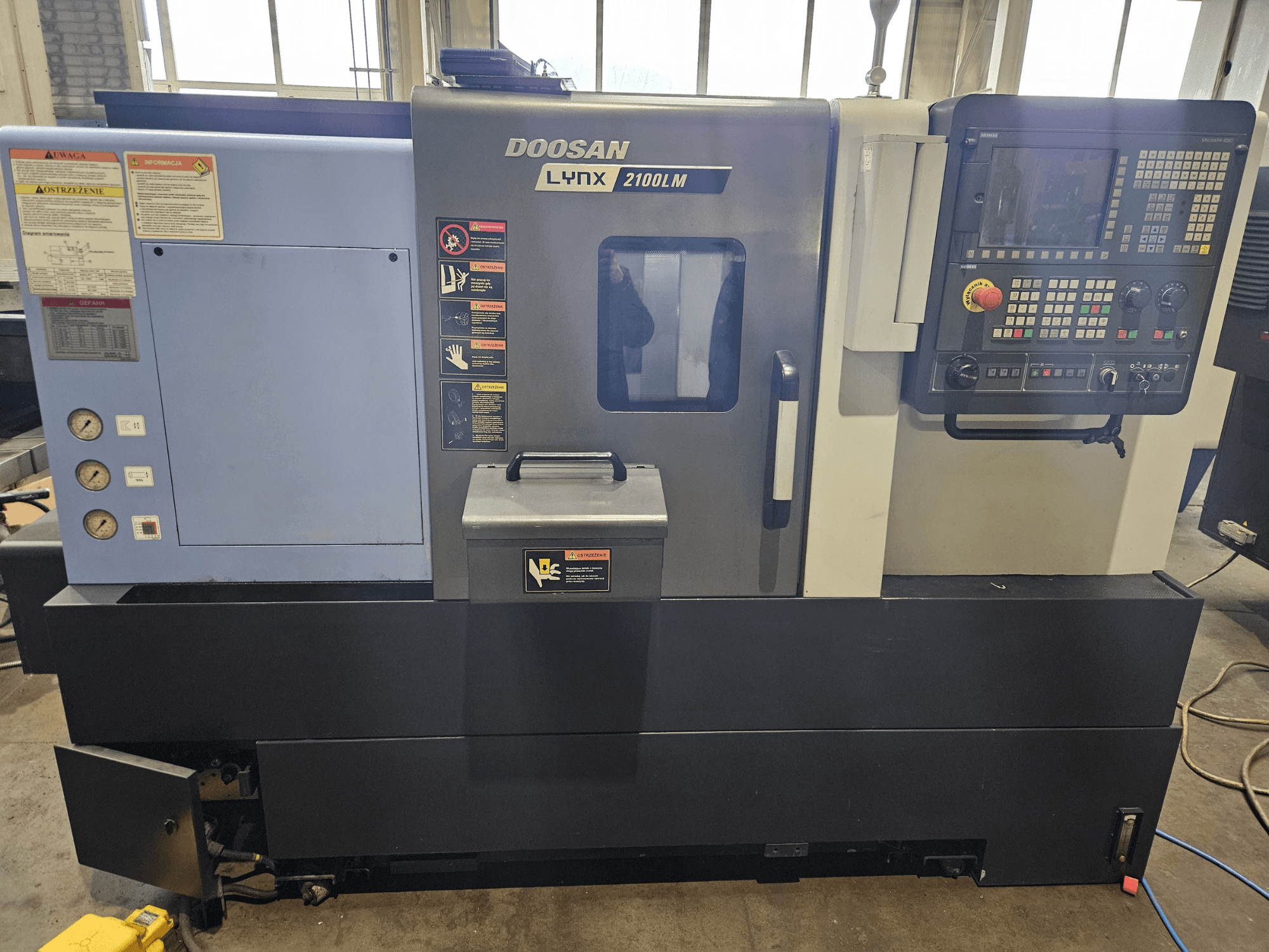 Doosan Lynx 2100LM CNC-Drehmaschine, Vorderansicht, mit Bedienfeld, Luftdruckmanometern und Sicherheitsschildern sichtbar.