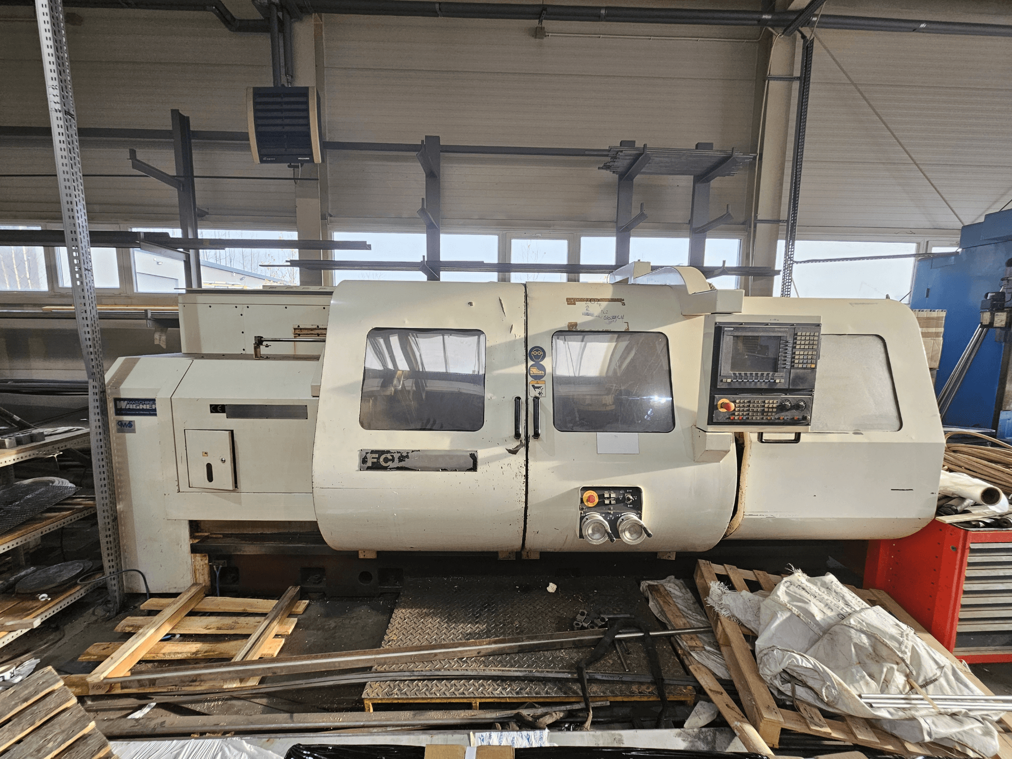 Wabeco CNC lathe, side view, featuring control panel, windows, and work area; machinery on wooden pallets in workshop setting.