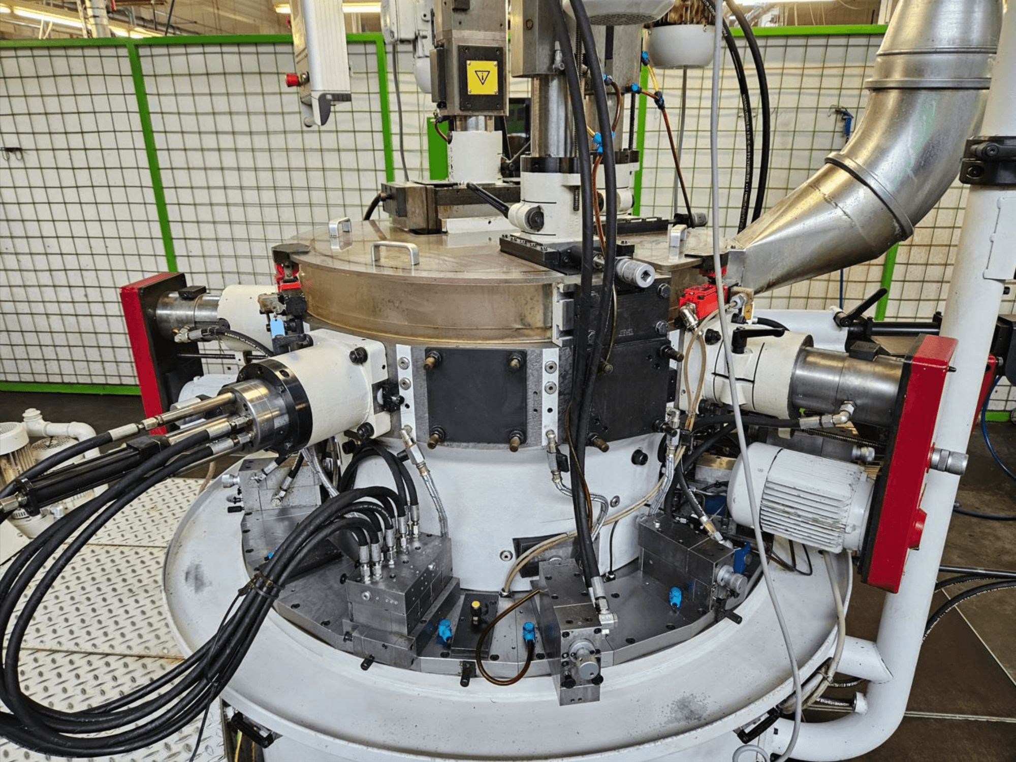 Nahaufnahme einer Industriemaschine mit Hydraulikschläuchen, Metallteilen und einem drehbaren Sockel. Keine Marke sichtbar.