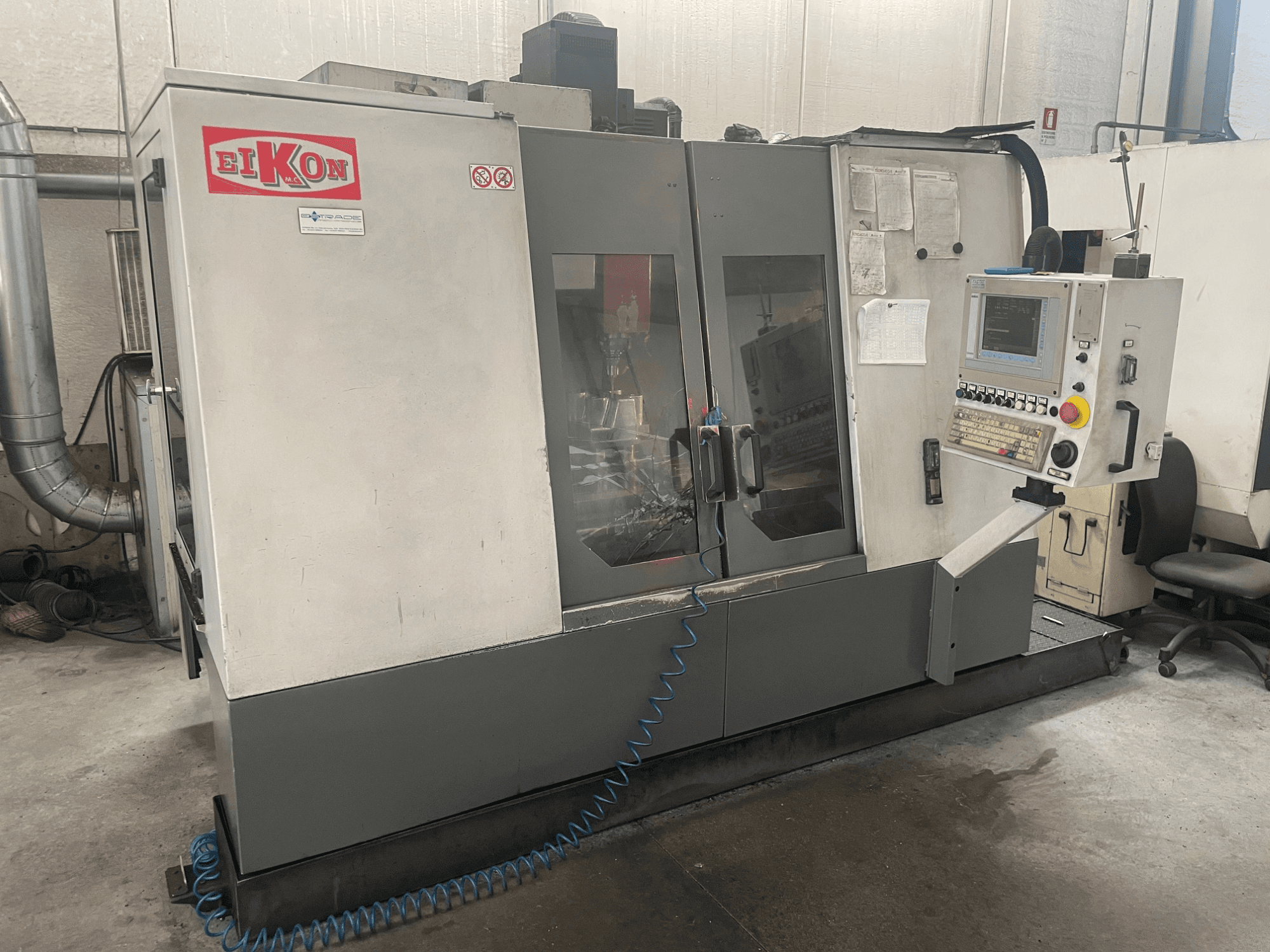 EIKON CNC-Maschine von vorne gesehen, mit Bedienfeld, großen Glastüren und einem blauen Luftschlauch auf dem Boden.