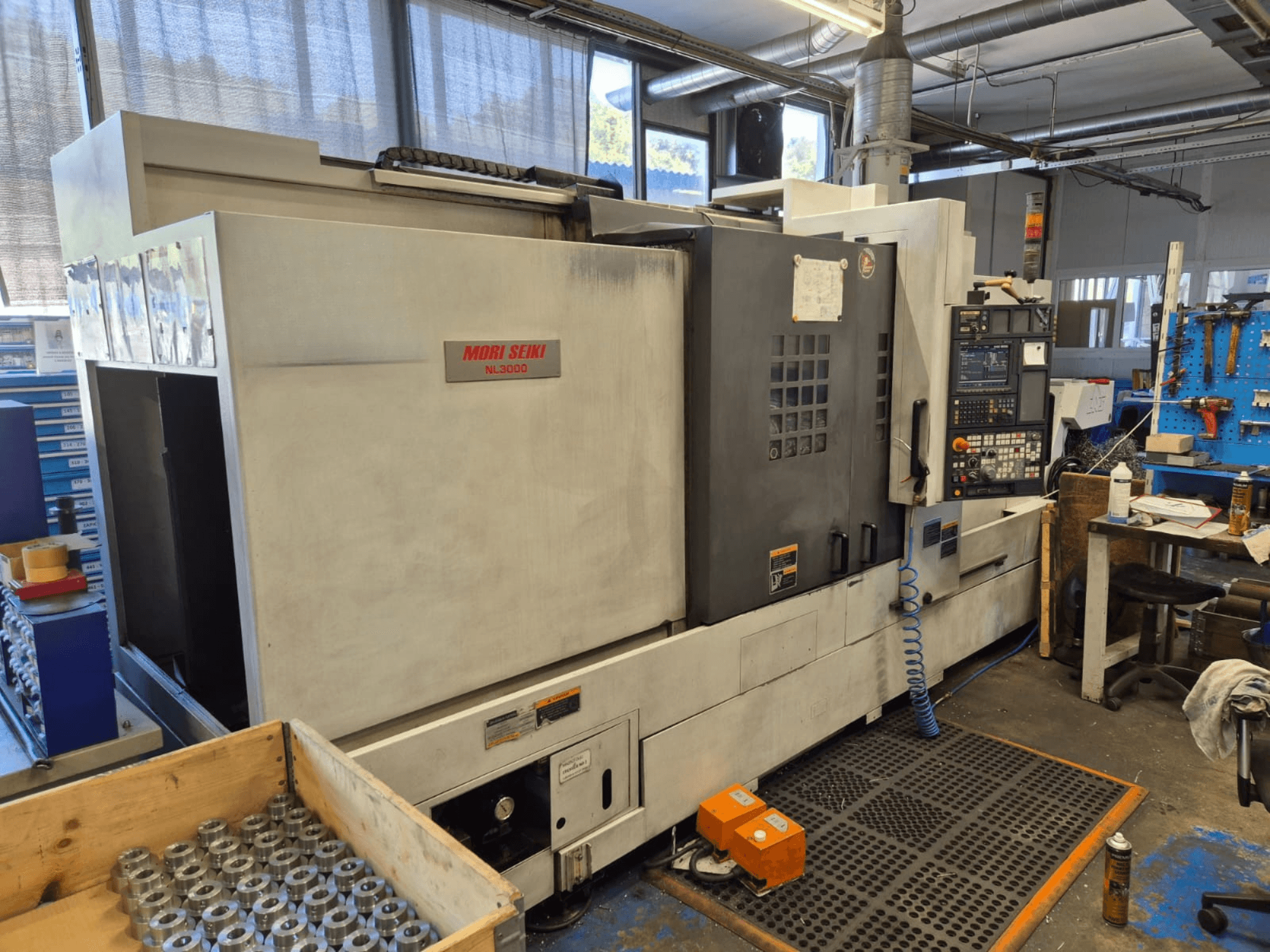 Mori Seiki NL3000 CNC-Drehmaschine in einer Werkstatt, Seitenansicht mit Bedienfeld, Werkzeugen und Metallteilen in einer Holzkiste.
