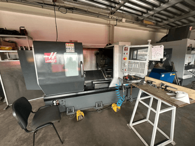 Haas CNC-Drehmaschine (Modell ST-10) in einer Werkstatt, Seitenansicht mit Bedienelementen, Werkzeugen auf einer Werkbank und einem Stuhl in der Nähe.