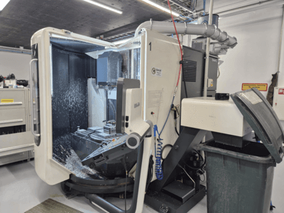 Geöffnete DMG-Fräsmaschine mit Kühlmittelnebel, mit Metallspänen und Werkzeugkontrollen, von vorne gesehen.