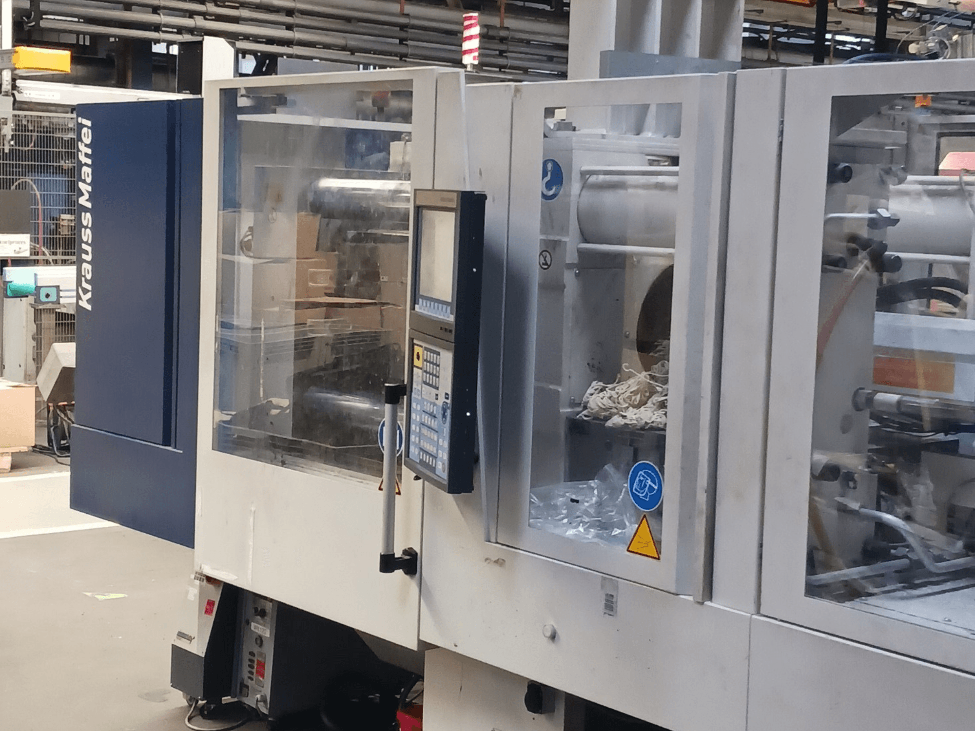 Spritzgießmaschine von KraussMaffei, Seitenansicht, mit Bedienfeld, Maschinen und Verpackungsmaterial im Hintergrund.
