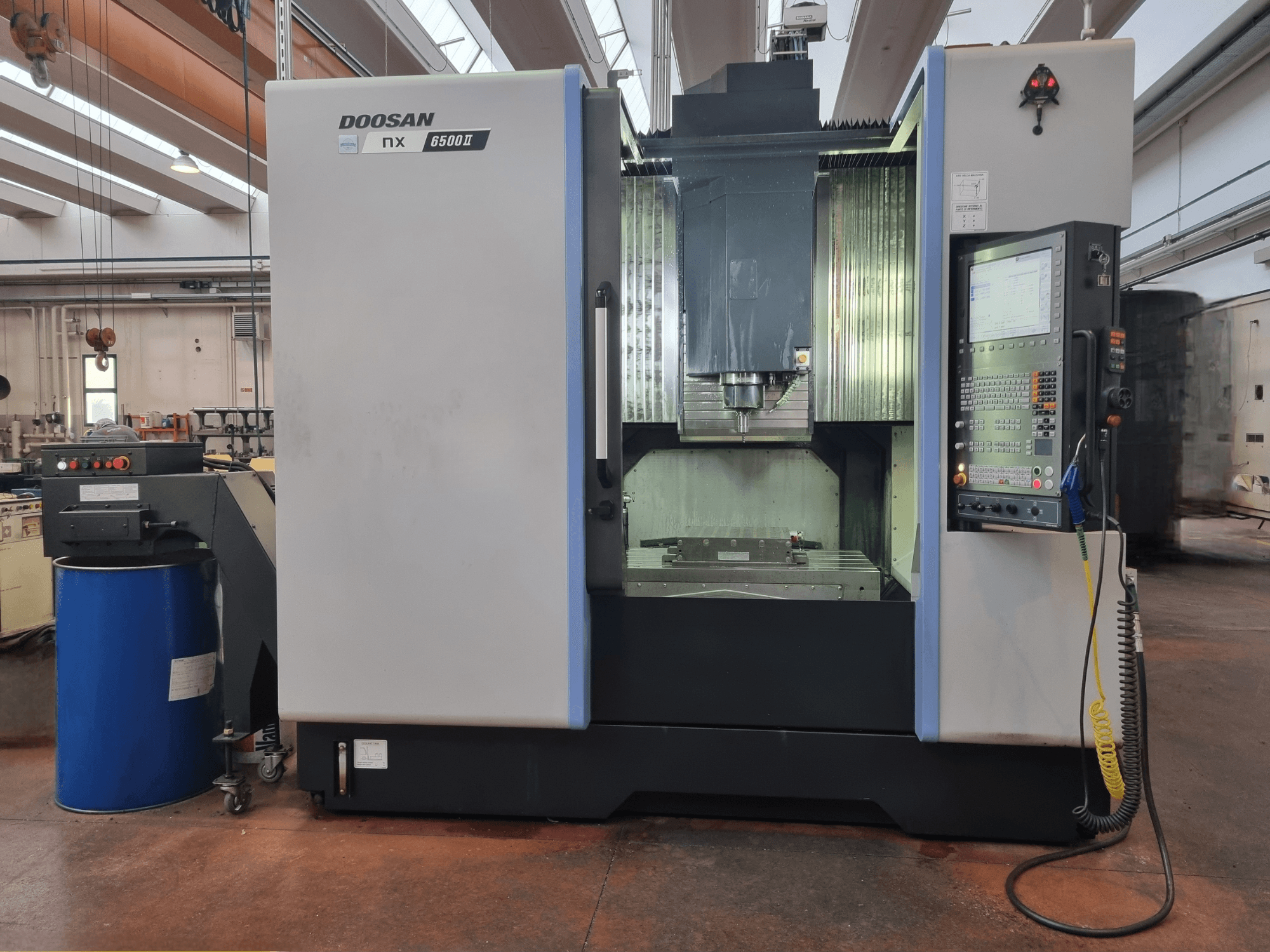 Doosan NX 6500 II CNC-Maschine, Vorderansicht, mit Bedienfeld und Bearbeitungsbereich, neben einer blauen Tonne positioniert.