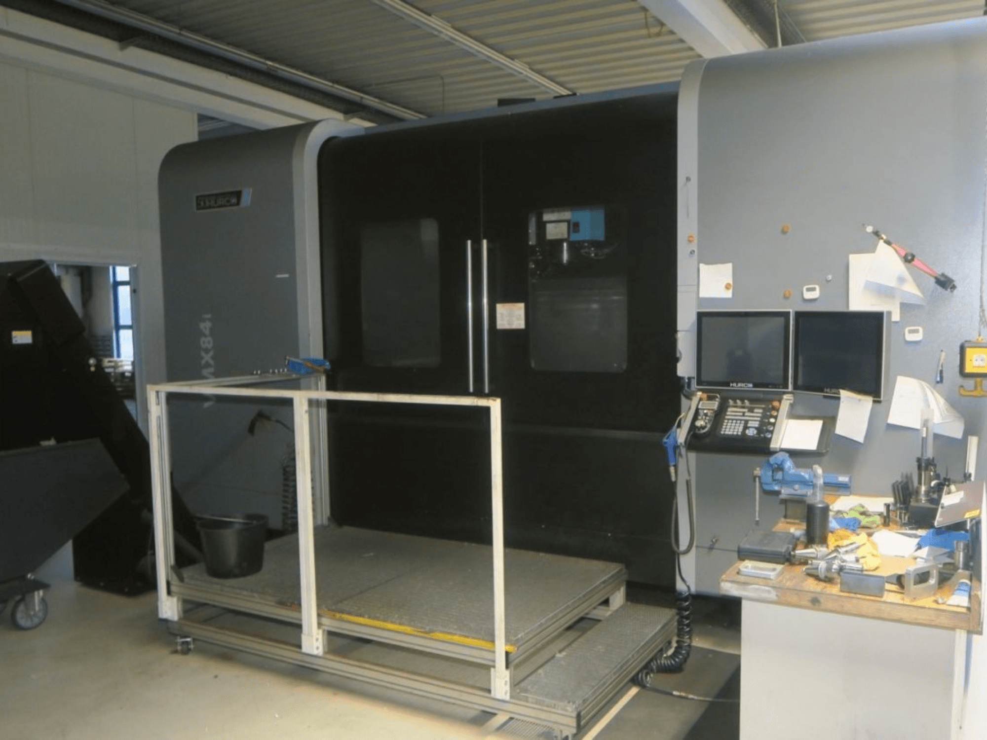 Hurco MX84 CNC-Maschine in der Vorderansicht, mit zwei Monitoren, Bedienfeld und Werkzeugarbeitsplatz mit verschiedenen Werkzeugen.