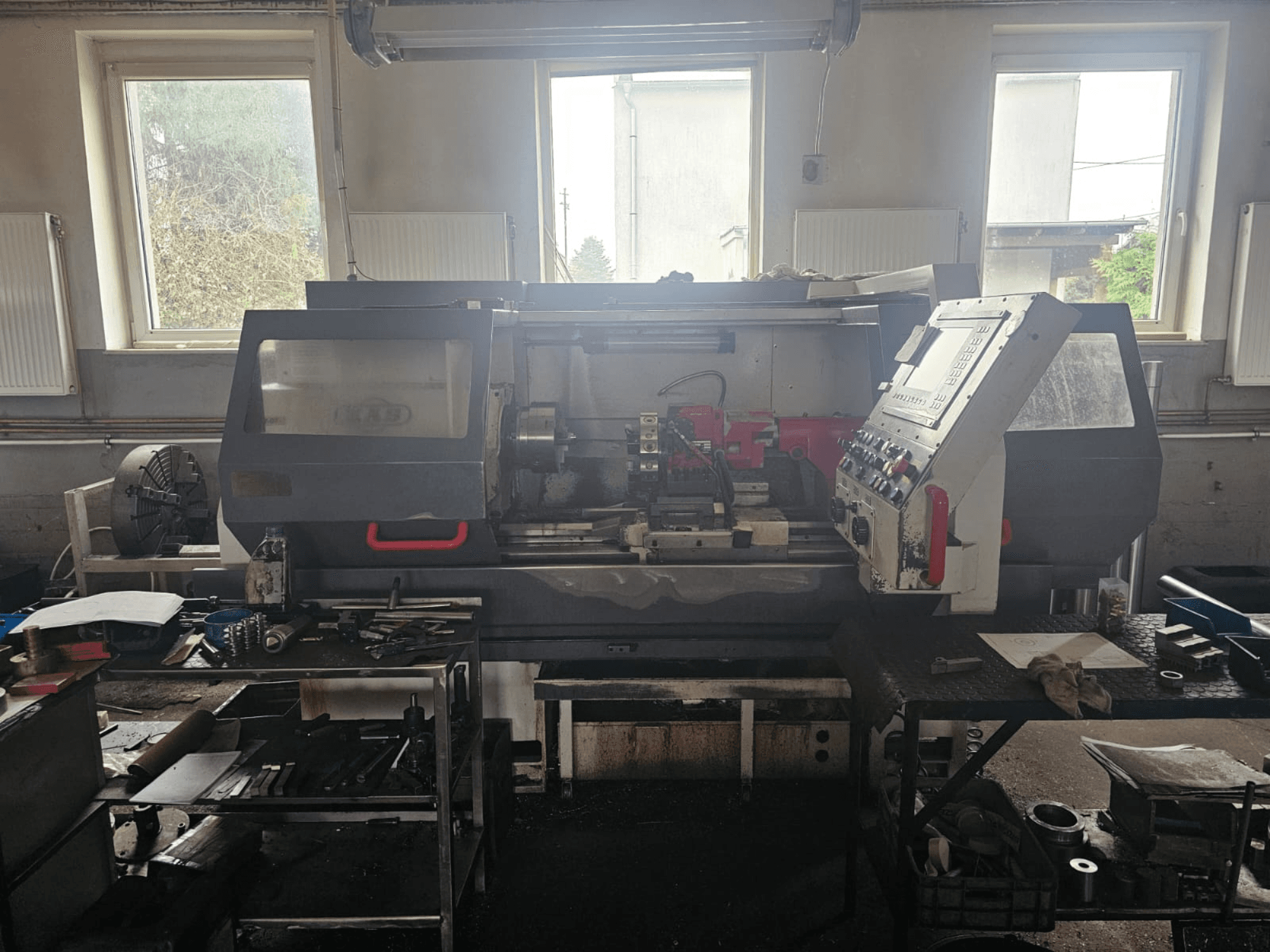 Drehmaschine in einer Werkstatt, Vorderansicht, mit Bedienfeld und Werkzeugen auf Tischen, umgeben von Fenstern und einem unübersichtlichen Arbeitsbereich.