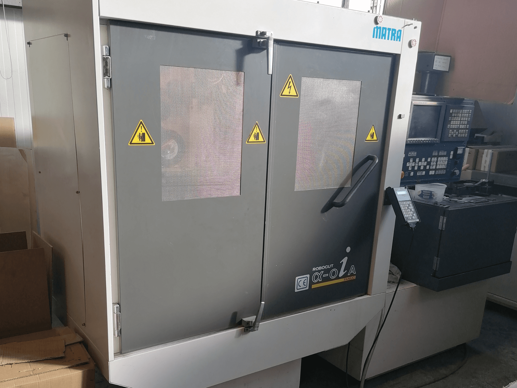 Robocut α-oZα-Maschine von Matra, Vorderansicht mit Sicherheitswarnungen, Bedienfeld und Doppeltüren mit Netzfenstern.