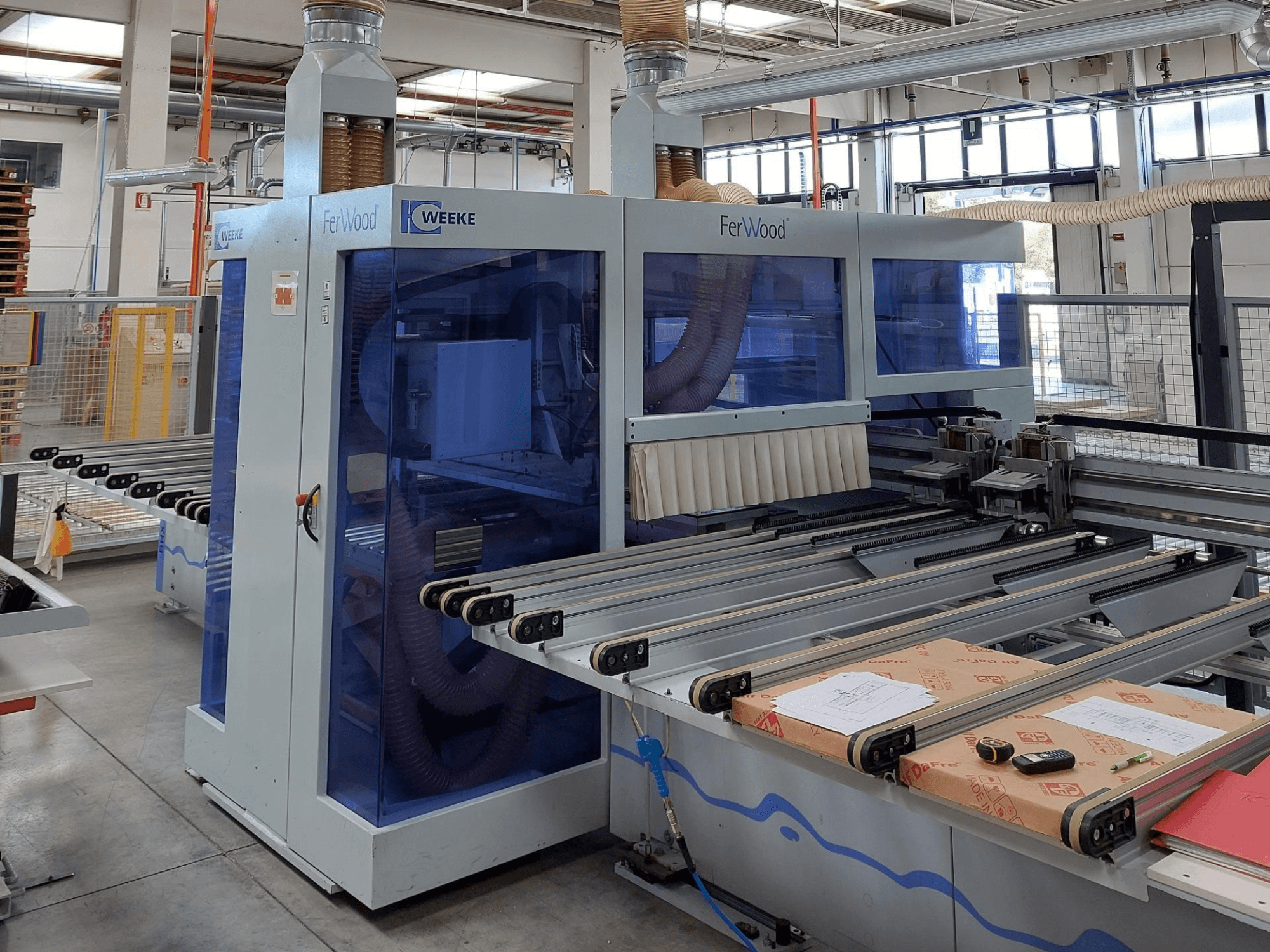 Weeke FerWood CNC-Maschine in einer Werkstatt, Vorderansicht mit blau getönten Bedienfeldern, Steuertasten und Arbeitstisch mit Materialien.