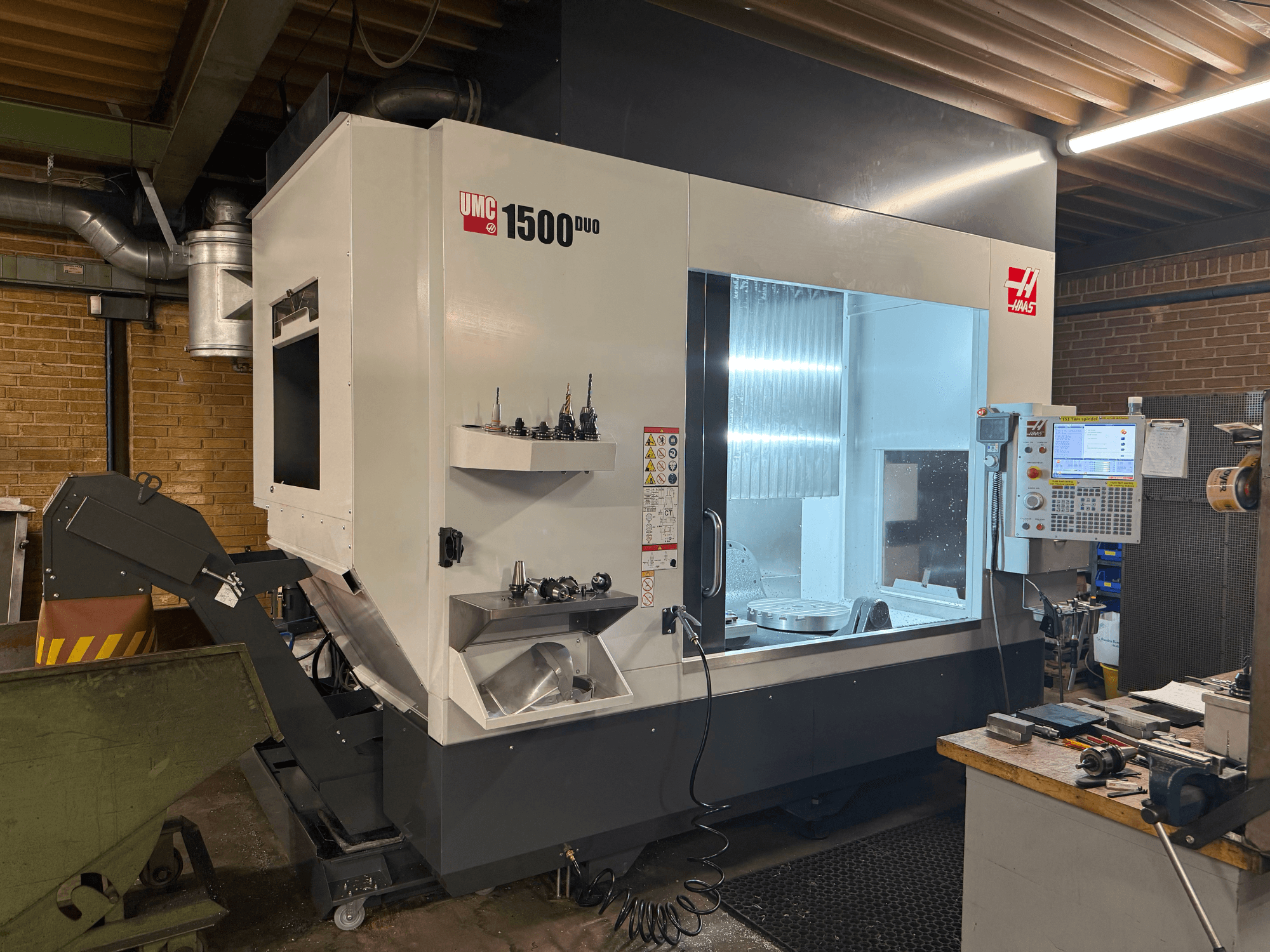 Haas UMC 1500DUO CNC-Maschine, Vorderansicht, mit einem großen Fenster, Bedienfeld und Werkzeughaltern auf einem Regal.