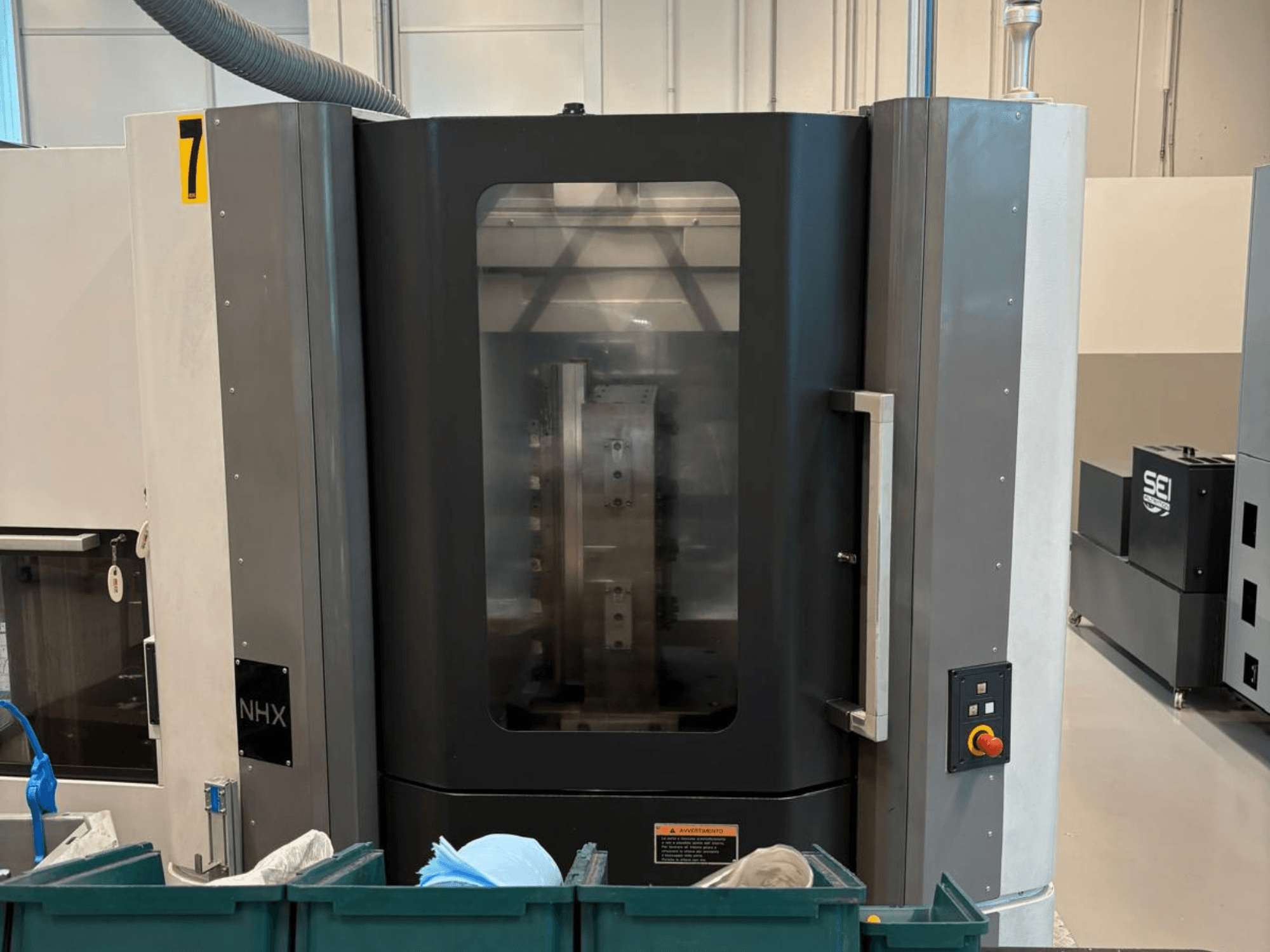 NHX-CNC-Maschine in der Vorderansicht mit Bedienfeld und übersichtlichem Display; sie steht in einer Werkstatt mit grünen Behältern in der Nähe.