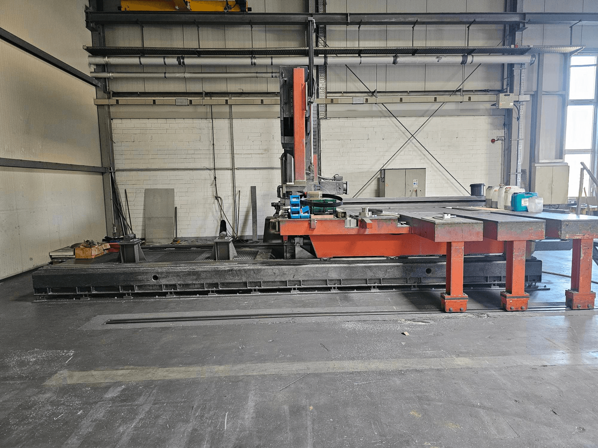 Industrial cutting machine in an open workshop, orange and black with a large table; side view showing accessories and workspace.