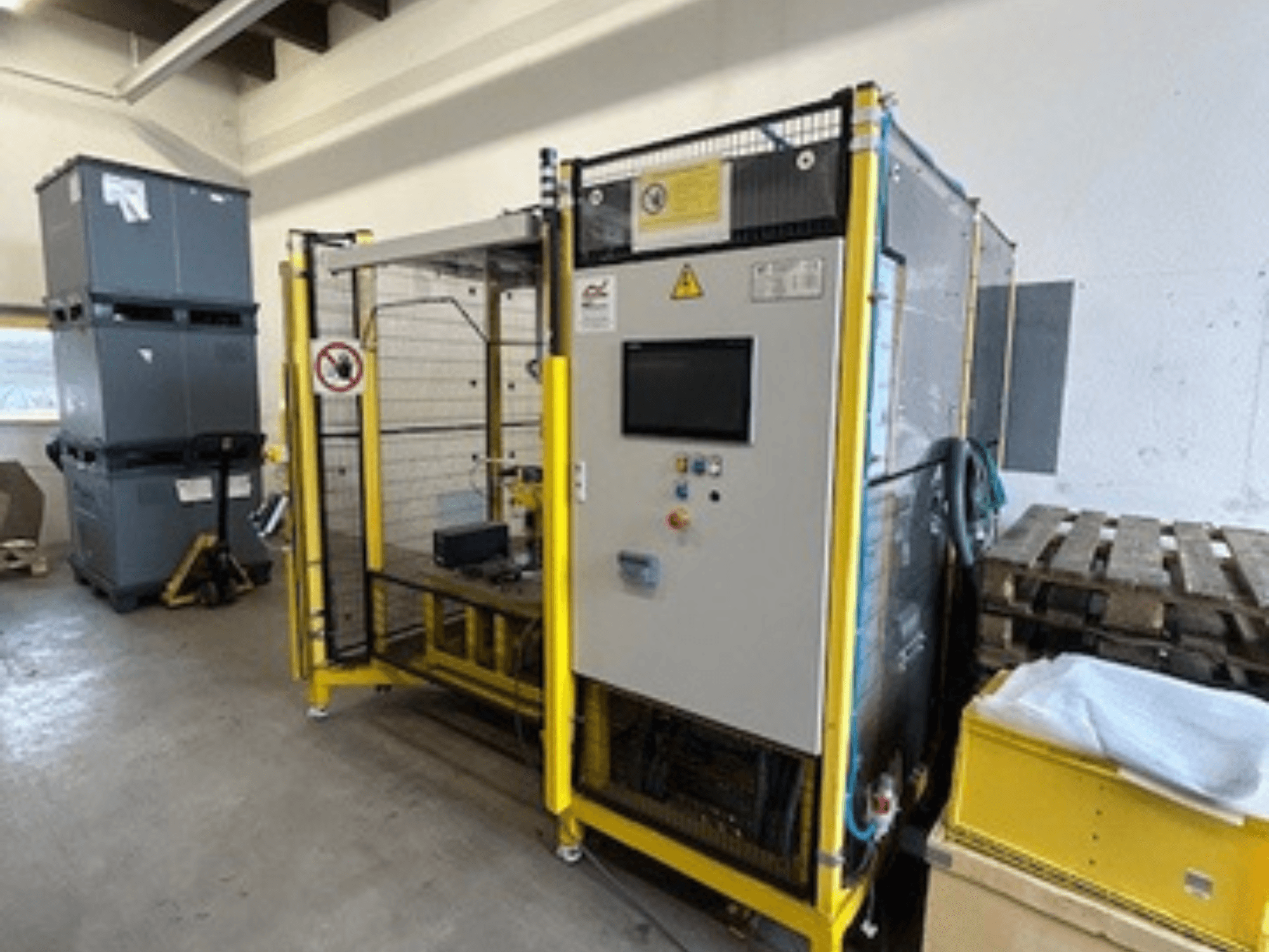 Industriemaschine mit einem Bedienfeld und Sicherheitseinrichtungen, Vorderansicht; gelber Rahmen, graue Behälter und Paletten im Hintergrund.