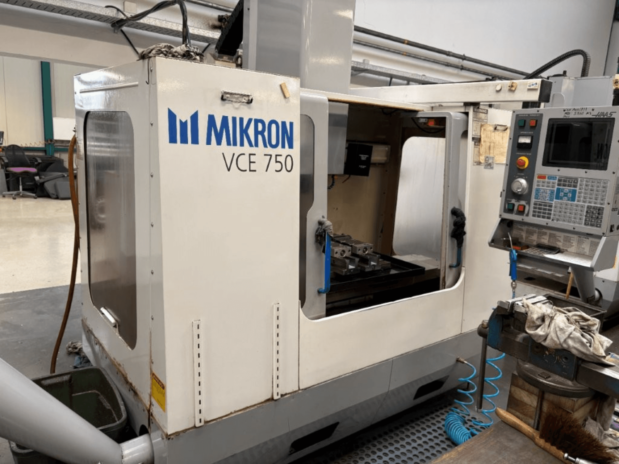Vorderansicht der Mikron VCE 750 CNC-Fräsmaschine mit einem bearbeiteten Arbeitstisch und einem Bedienfeld mit verschiedenen Tasten und Bildschirmen.