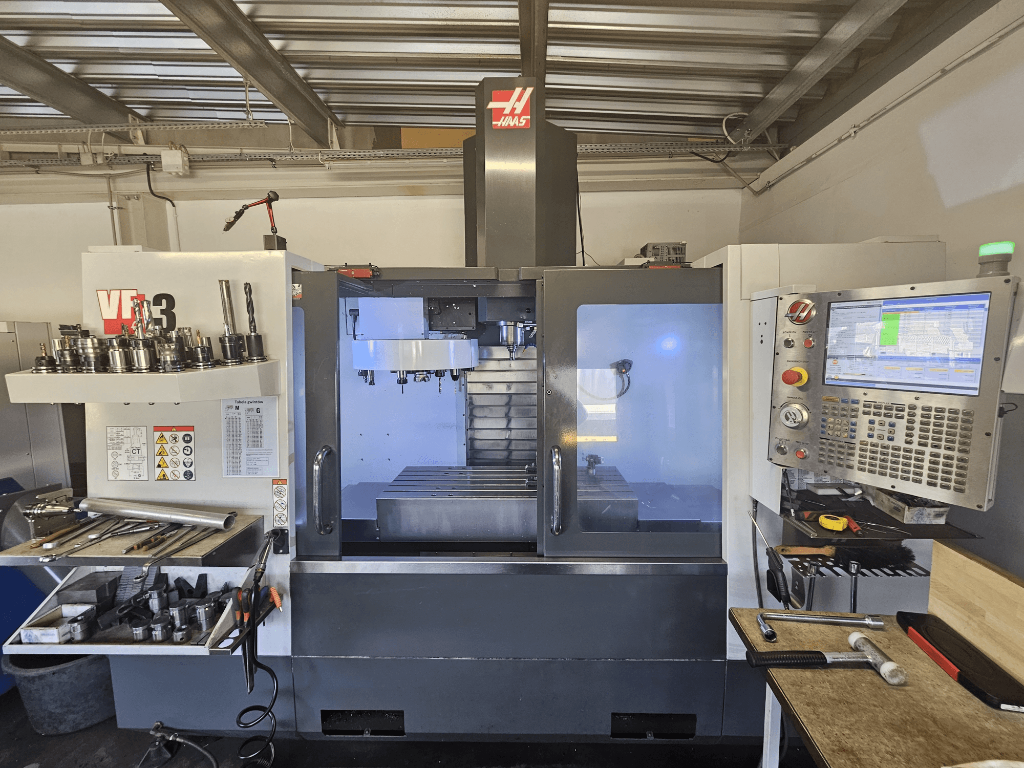 Haas VF-3 CNC-Maschine in einer Werkstatt, Vorderansicht mit Werkzeugen, Bedienelementen und Fräsbereich mit beleuchtetem Innenraum.