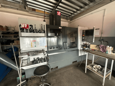 Haas VF-3 CNC-Maschine in der Werkstatt, Vorderansicht mit Werkzeugen und Bedienelementen auf dem Nebentisch, Anzeige der technischen Schnittstelle und Sicherheitsinformationen.