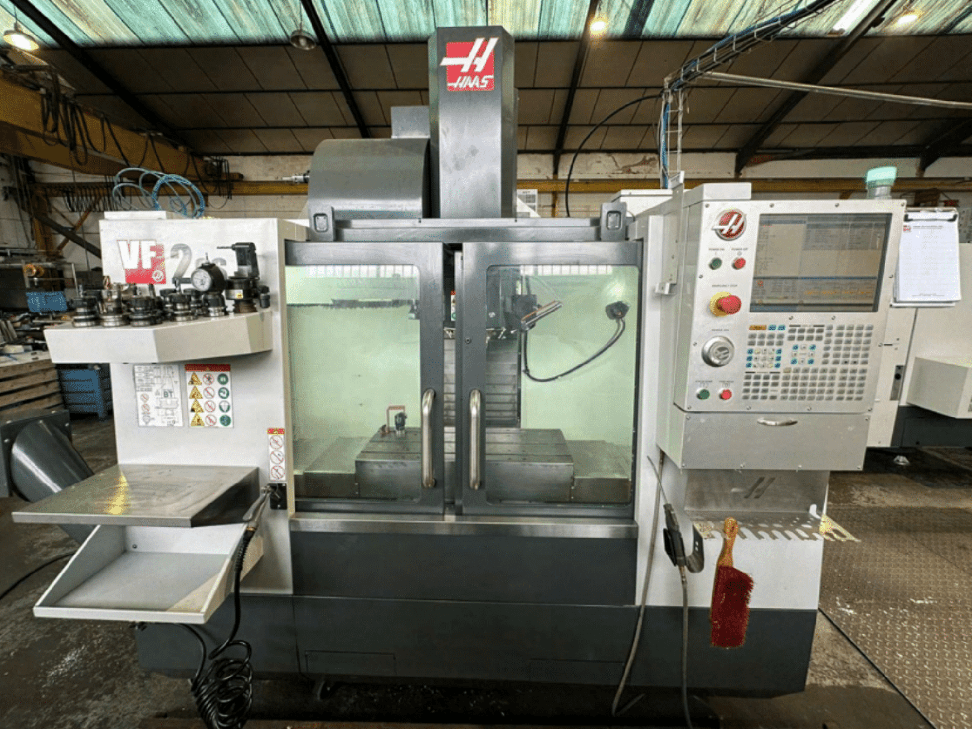 Haas VF2 CNC machine front view, showing control panel, tool holders, and workspace with safety labels and a brush.