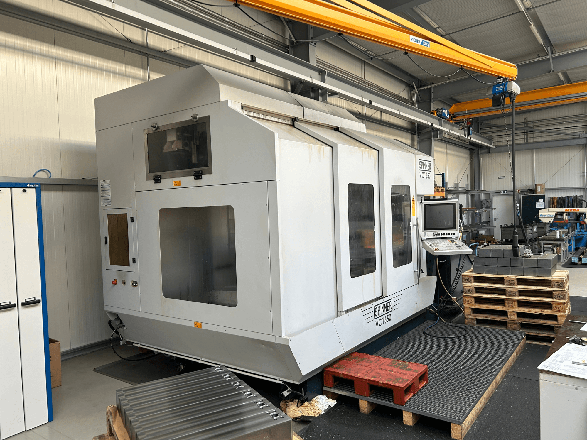 Spinner VC 1650 CNC-Maschine, Vorderansicht, mit Bedienfeld, großen Fenstern und in der Nähe gestapelten Paletten in einer Werkstattumgebung.