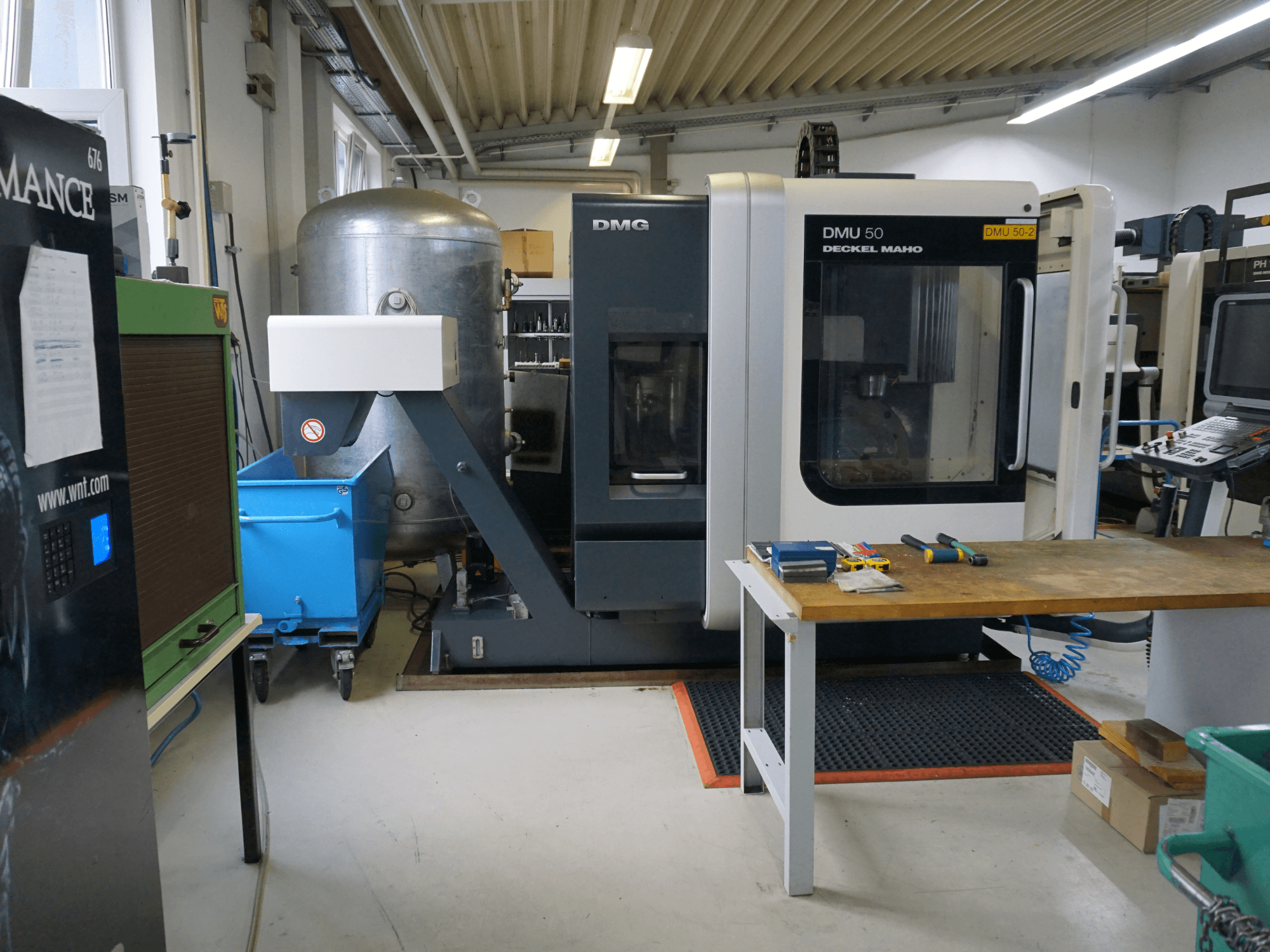 DMG DMU 50 milling machine in a workshop, front view, with tools on a wooden table and a blue wheeled bin nearby.
