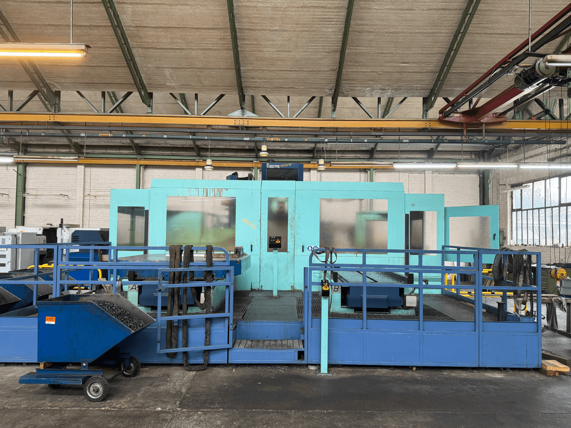Industrial blue CNC machine with protective panels, front view, featuring tool storage and a waste collection cart on the side.