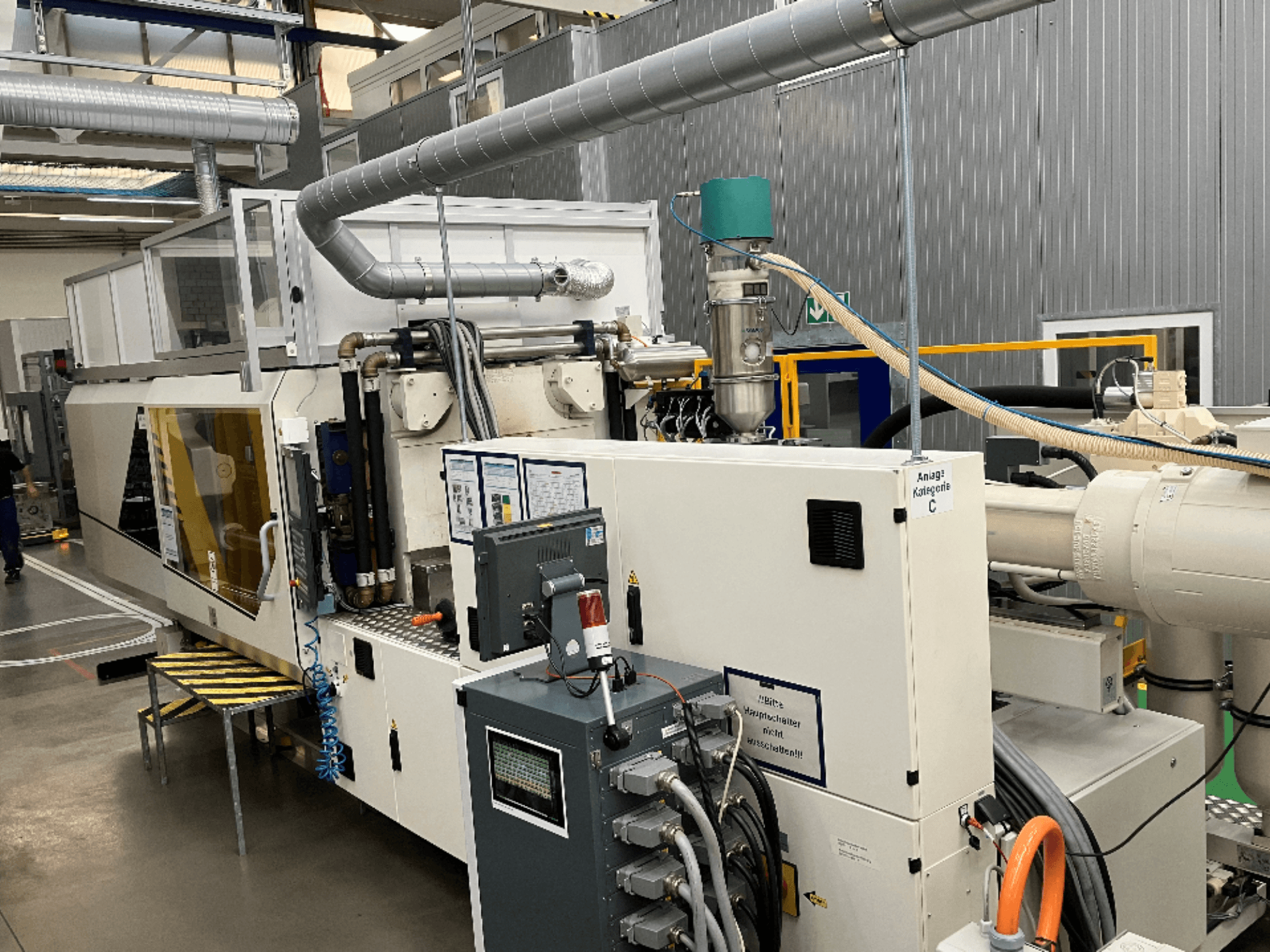 Injection molding machine, front view, featuring brand label, control panel, and visible hoses in a factory setting.