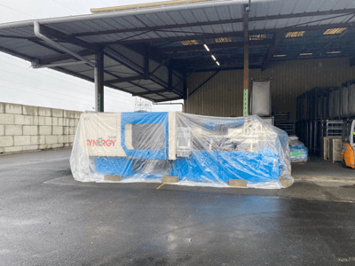 Verpackte SYNERGY 2400 Maschine von vorne gesehen, blaue und beige Farben, innerhalb einer großen überdachten Industriefläche.