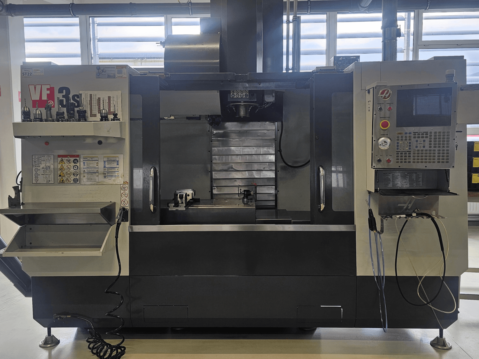 Haas VF3SS CNC-Fräse, Vorderansicht, mit Werkzeughaltern, Bedienfeld und leerer Kühlmittelwanne, mit sichtbaren Sicherheitsschildern.