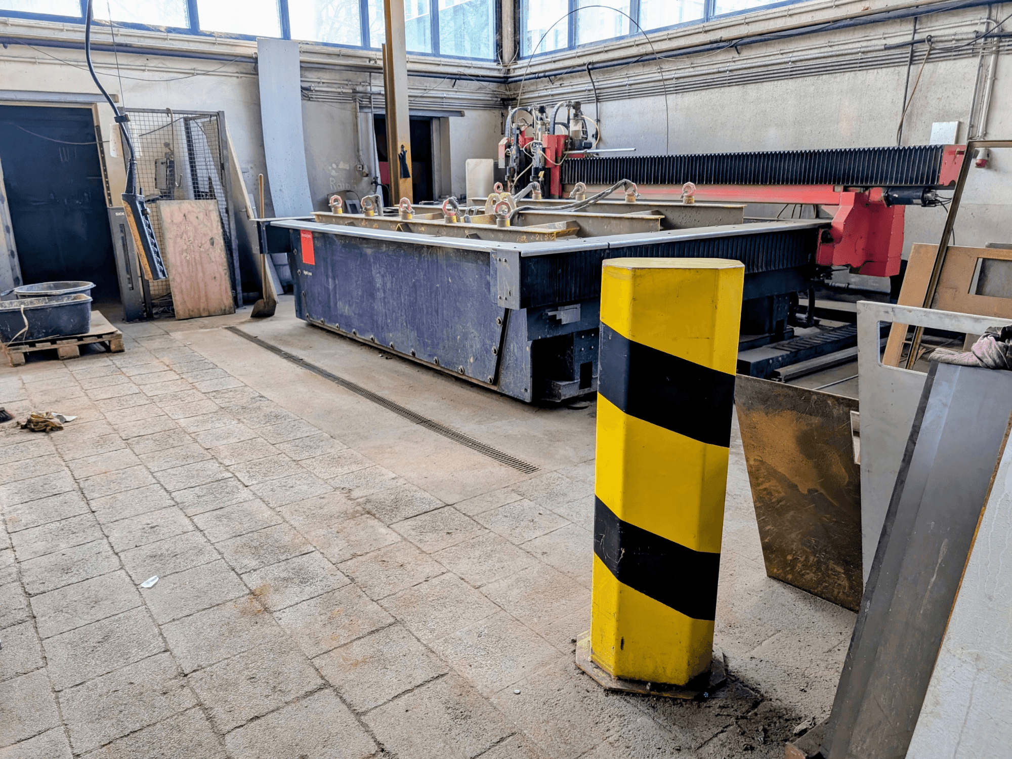 Bystronic Byjet L 2030 Wasserstrahlschneidanlage in einer Werkstatt, Seitenansicht, mit gelber Sicherheitsstange und umliegenden Materialien.