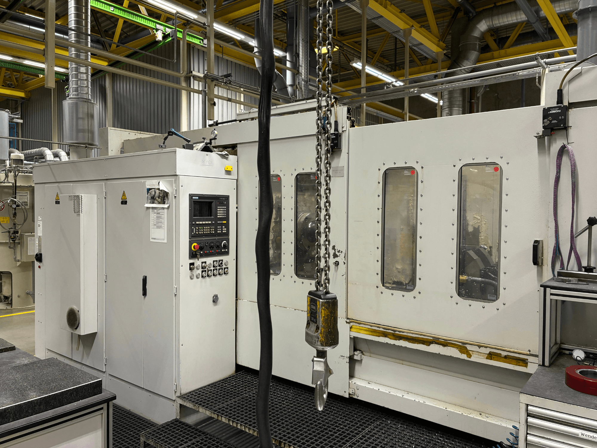 Industrielle CNC-Maschine mit Bedienfeld, Seitenansicht; mit Fenstern, Ketten und Metallkomponenten in einer Fabrikumgebung.