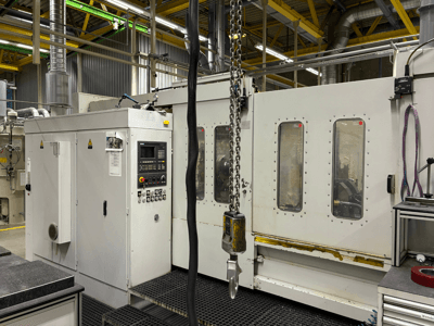 Industrielle CNC-Maschine mit Bedienfeld, Seitenansicht; mit Fenstern, Ketten und Metallkomponenten in einer Fabrikumgebung.